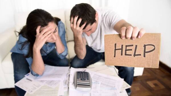 Man holding up help sign with woman with her head in hands. A calculator and bills lay on the table